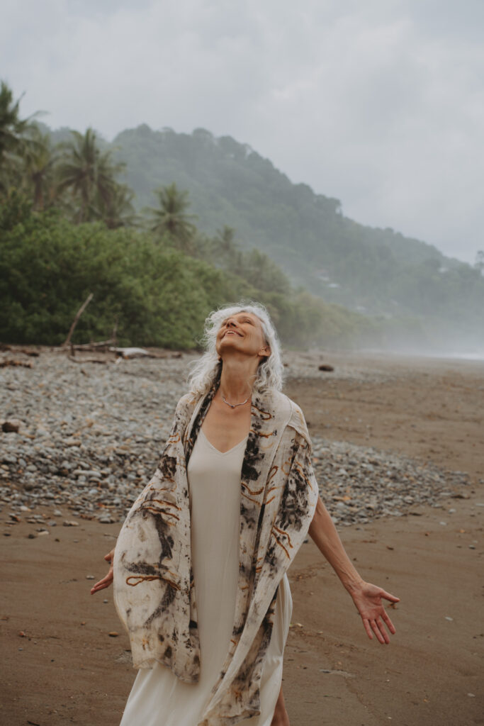 A woman with long gray hair stands on a rocky beach, eyes closed and arms outstretched, wearing a light dress and patterned shawl. Behind her, there are palm trees and misty hills under a cloudy sky.