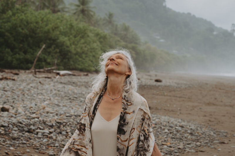 A woman with long gray hair stands on a rocky beach, eyes closed and arms outstretched, wearing a light dress and patterned shawl. Behind her, there are palm trees and misty hills under a cloudy sky.