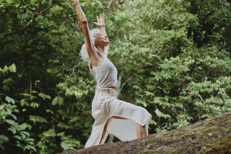 A person in a white dress performs a yoga pose on a large rock surrounded by lush green foliage. Their arms are stretched upward, and they are barefoot, conveying a sense of harmony with nature.