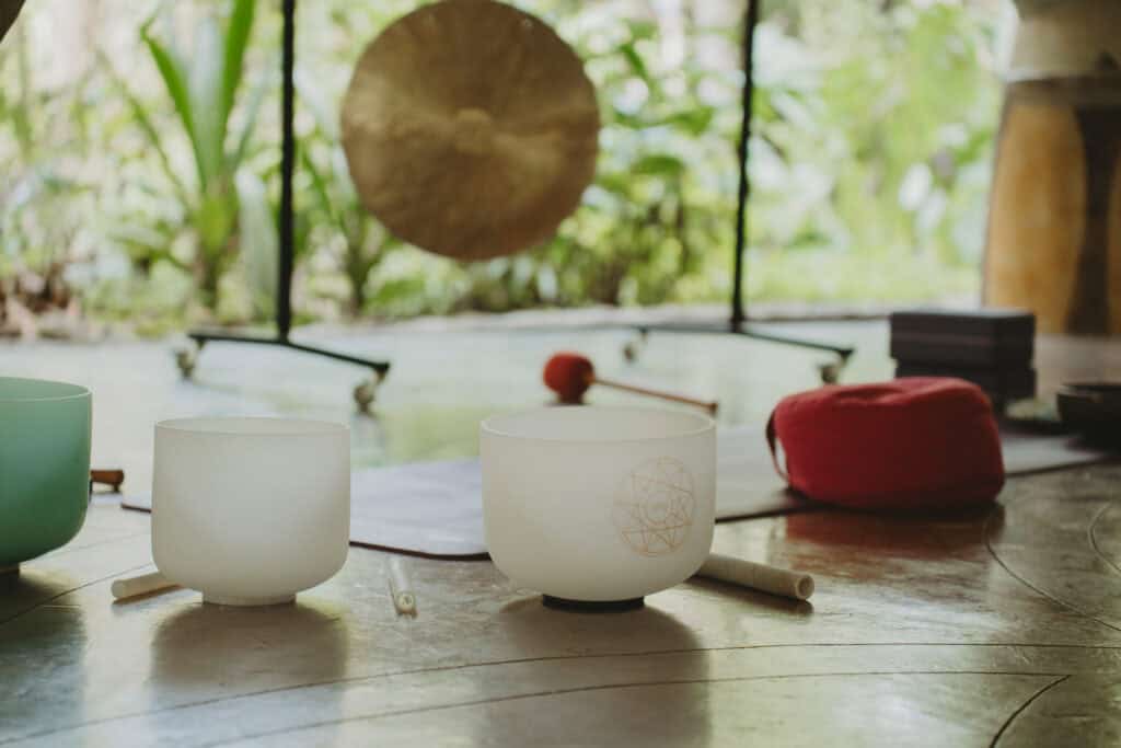 White crystal singing bowls and mallets are arranged on a wooden floor in a peaceful, green indoor setting. In the background, a gong and yoga mats add to the serene atmosphere.