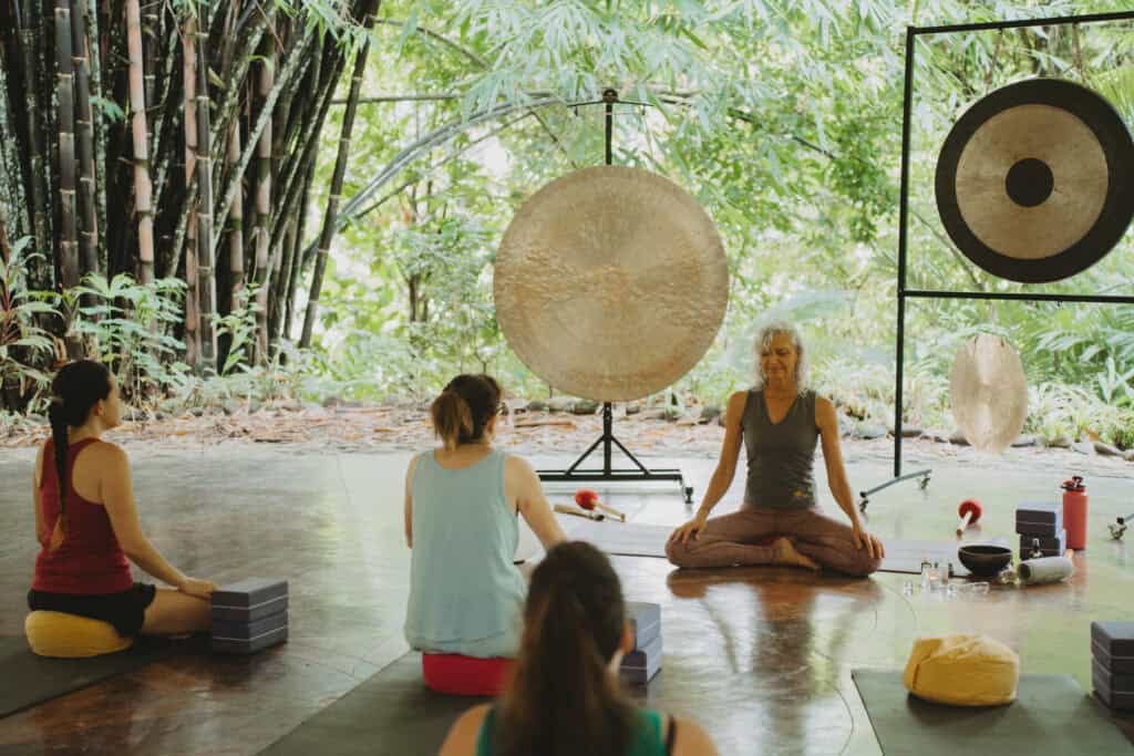 A woman sits cross-legged in front of three gongs, leading a group yoga session in a lush, forested setting. Four participants sit on mats facing her, surrounded by yoga blocks and greenery.