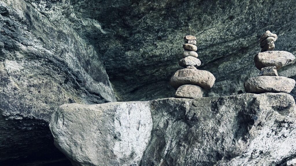 Two small towers of smooth stones are stacked on a large rock ledge beneath an overhanging rocky formation, creating a sense of balance and tranquility in a natural, rugged setting.