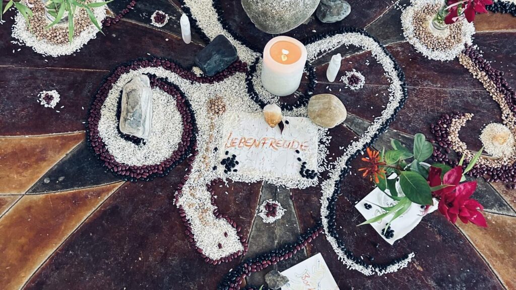 A decorative arrangement on a wooden surface features rice, beans, stones, flowers, candles, and a card with "LEBENFREUDE" written on it, creating a colorful and artistic display.