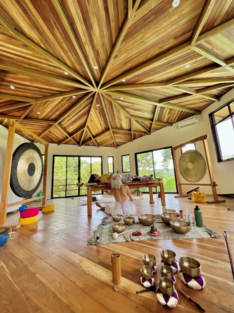 A person sits cross-legged on a mat in a spacious, wooden-ceilinged room with large windows. Gongs, singing bowls, and meditation tools are arranged around them, creating a peaceful, serene atmosphere.