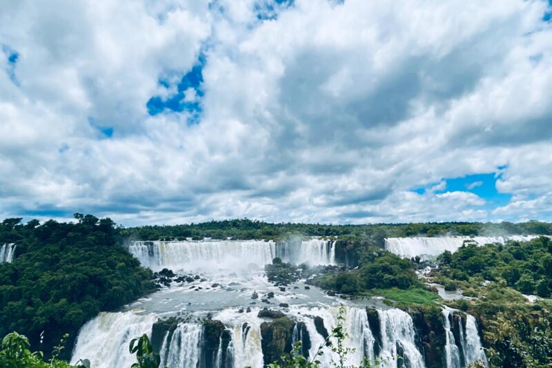 Waterfalls cascade over lush green cliffs under a partly cloudy blue sky, with vibrant greenery in the foreground and mist rising from the falls.