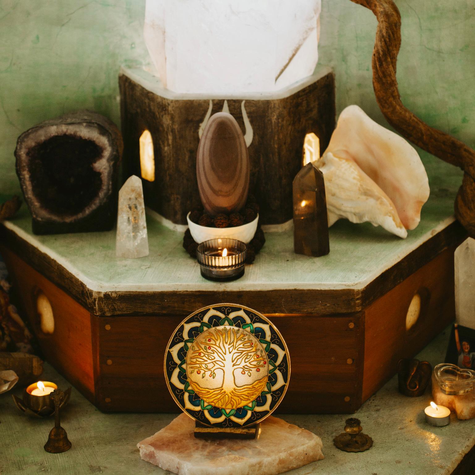 A spiritual altar with crystals, a geode, shells, candles, and a circular mandala featuring a golden tree, all arranged on a hexagonal wooden table against a green background.