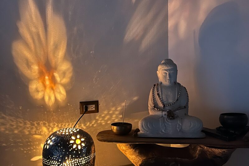 A peaceful corner with a Buddha statue on a wooden table, surrounded by candles and bowls. A glowing lamp with flower-shaped holes casts warm, intricate patterns on the wall, creating a serene, meditative atmosphere.
