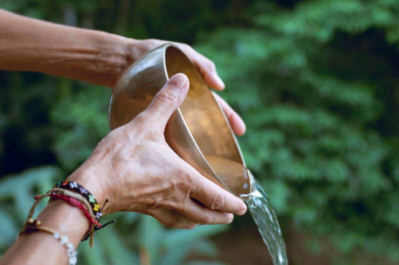 A person pours water from a metal bowl outdoors, with greenery and plants in the background. The person is wearing colorful bracelets on their wrist.
