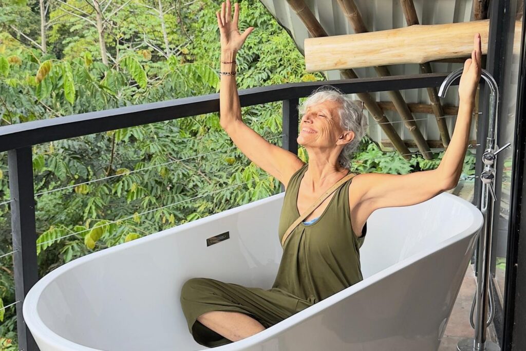 An older woman with gray hair, wearing a green sleeveless jumpsuit, sits playfully in an empty outdoor bathtub, smiling with both arms raised. Lush greenery and a metal railing are visible in the background.