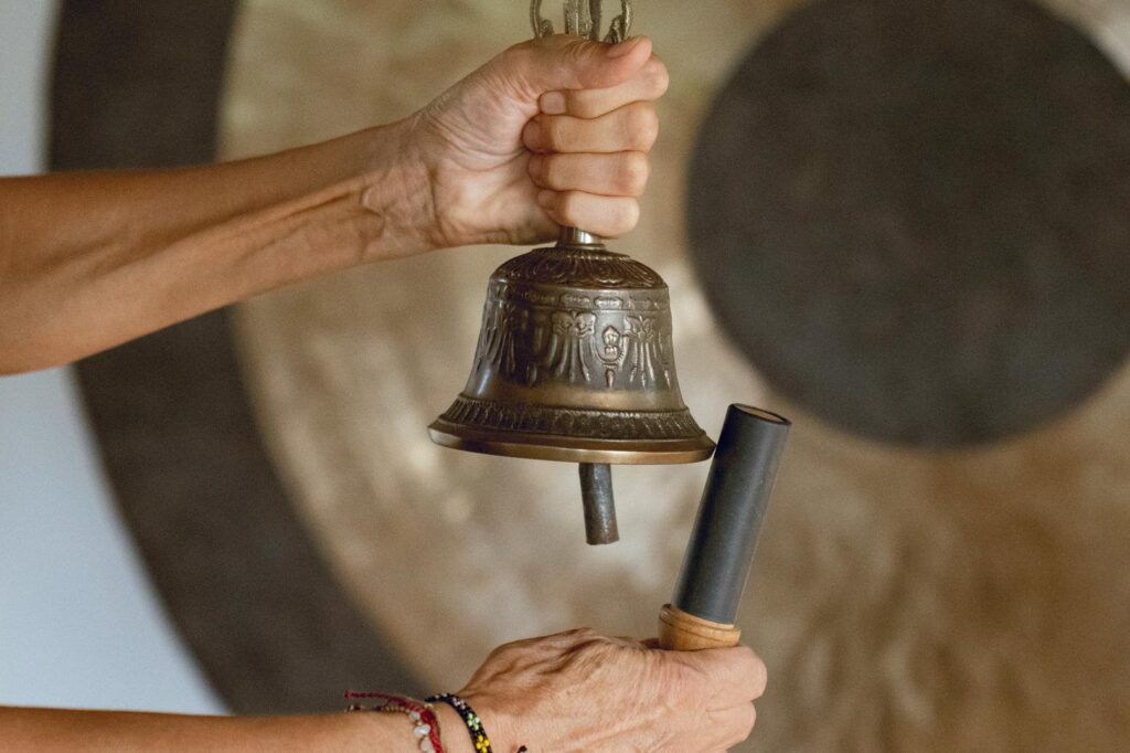 A person’s hands holding a decorative metal bell and a striker in front of a large gong, ready to strike the bell. The person wears a beaded bracelet on their wrist.