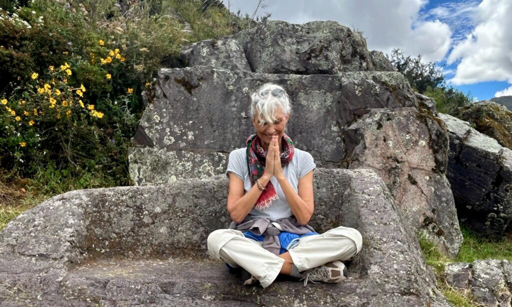 An older woman with short gray hair sits cross-legged on a stone bench outdoors, hands pressed together in front of her face, surrounded by rocks and greenery under a partly cloudy sky.