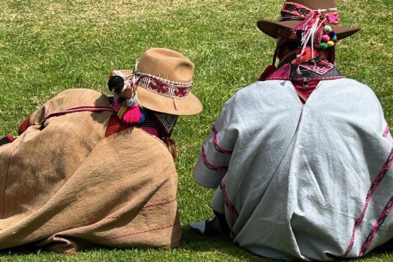 Two people sit on the grass wearing traditional Andean clothing, including tan and brown hats with decorative bands and colorful accents, seen from behind on a sunny day.