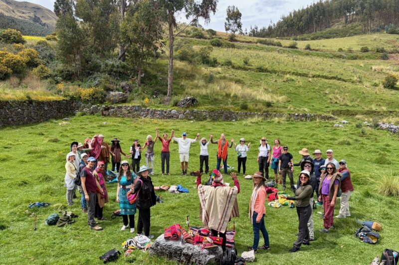 A group of people stand in a circle on a grassy field surrounded by trees and hills, holding hands and raising their arms in celebration or unity. Backpacks and belongings are scattered nearby.