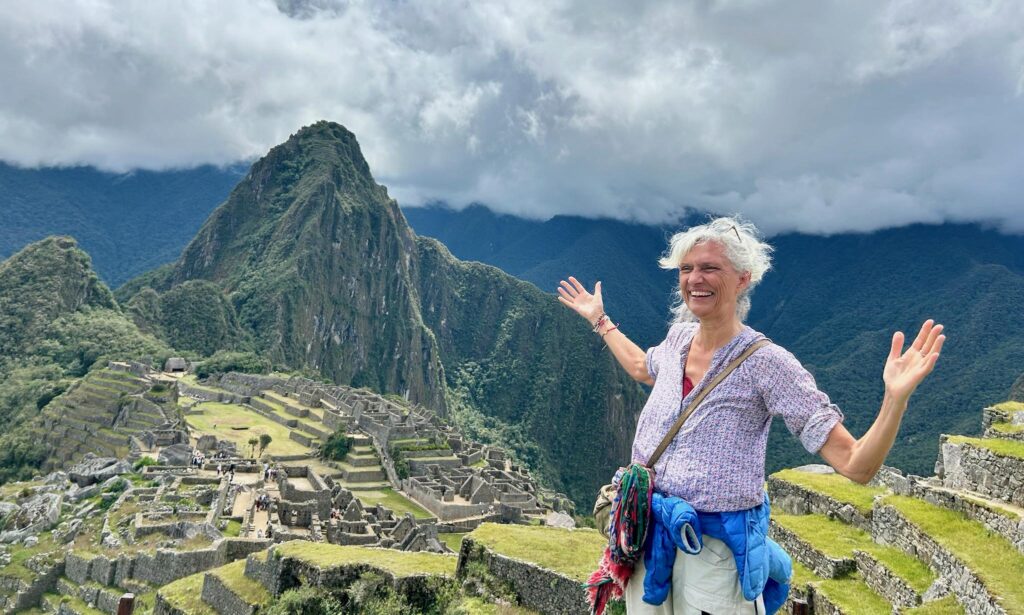 A smiling person with short gray hair stands with arms outstretched in front of the ancient ruins of Machu Picchu, surrounded by lush green mountains under a cloudy sky.