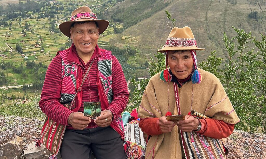 Two people wearing traditional Andean clothing and hats sit outdoors on a hillside, smiling and holding cards, with a green valley and mountains in the background.