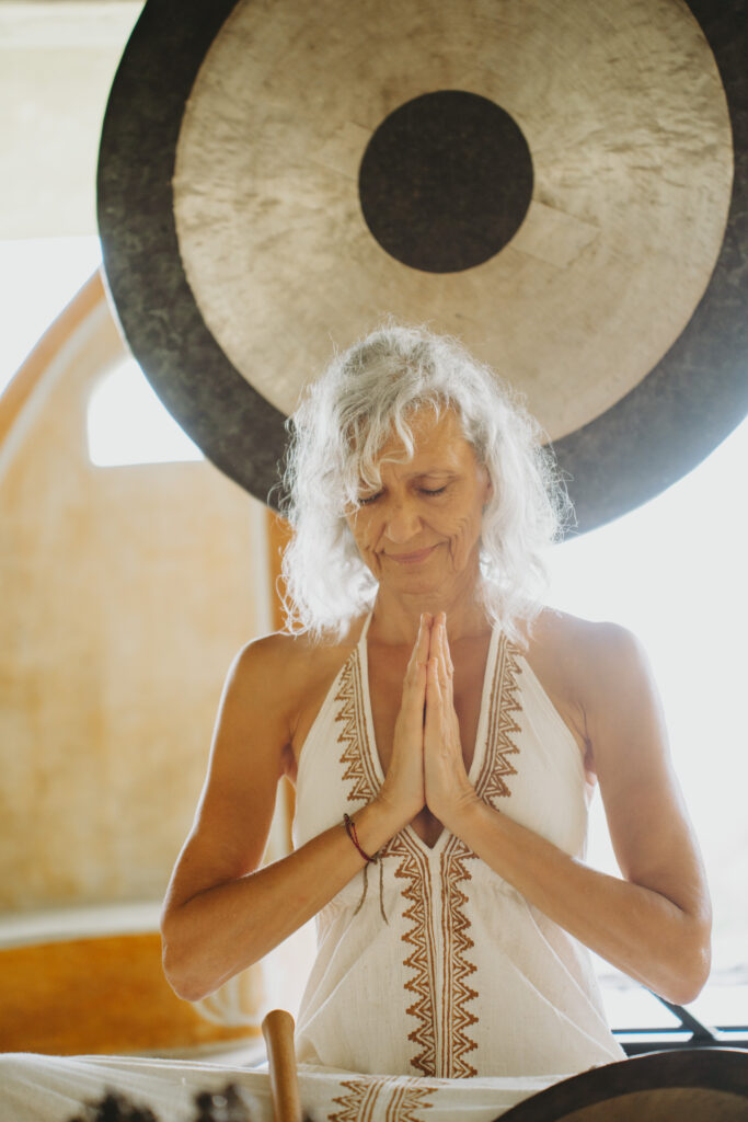 A woman with gray curly hair, wearing a white dress with embroidered details, stands with palms pressed together in front of a large gong, eyes closed, appearing peaceful and meditative in a softly lit room.