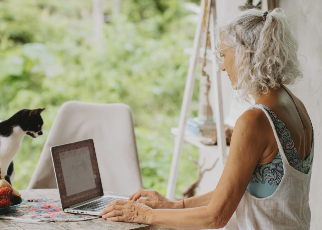 An older woman with gray hair uses a laptop at a rustic outdoor table, while a black and white cat sits nearby. Lush greenery is visible in the background. The setting appears calm and cozy.