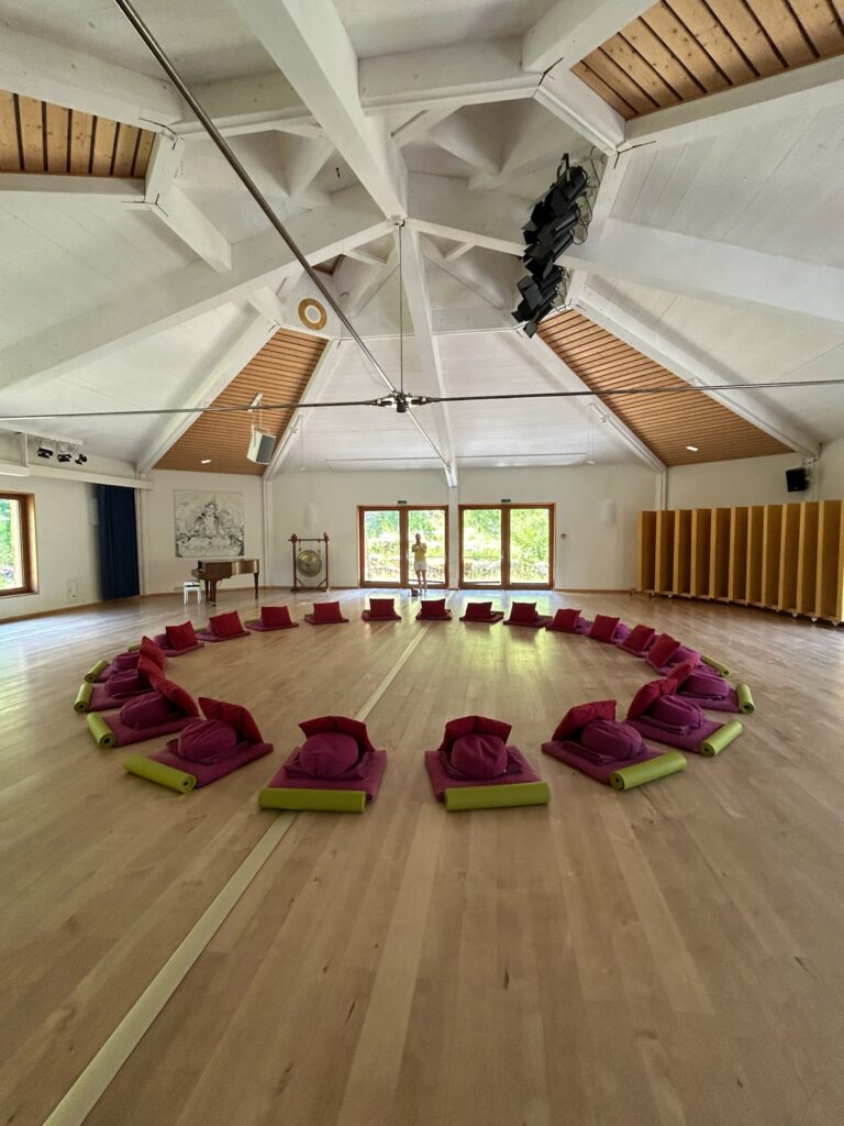 A spacious, bright room with a wooden ceiling has meditation cushions arranged in two circles on a light wood floor, facing inward toward the center. Large windows let in natural light, and shelves line the wall.