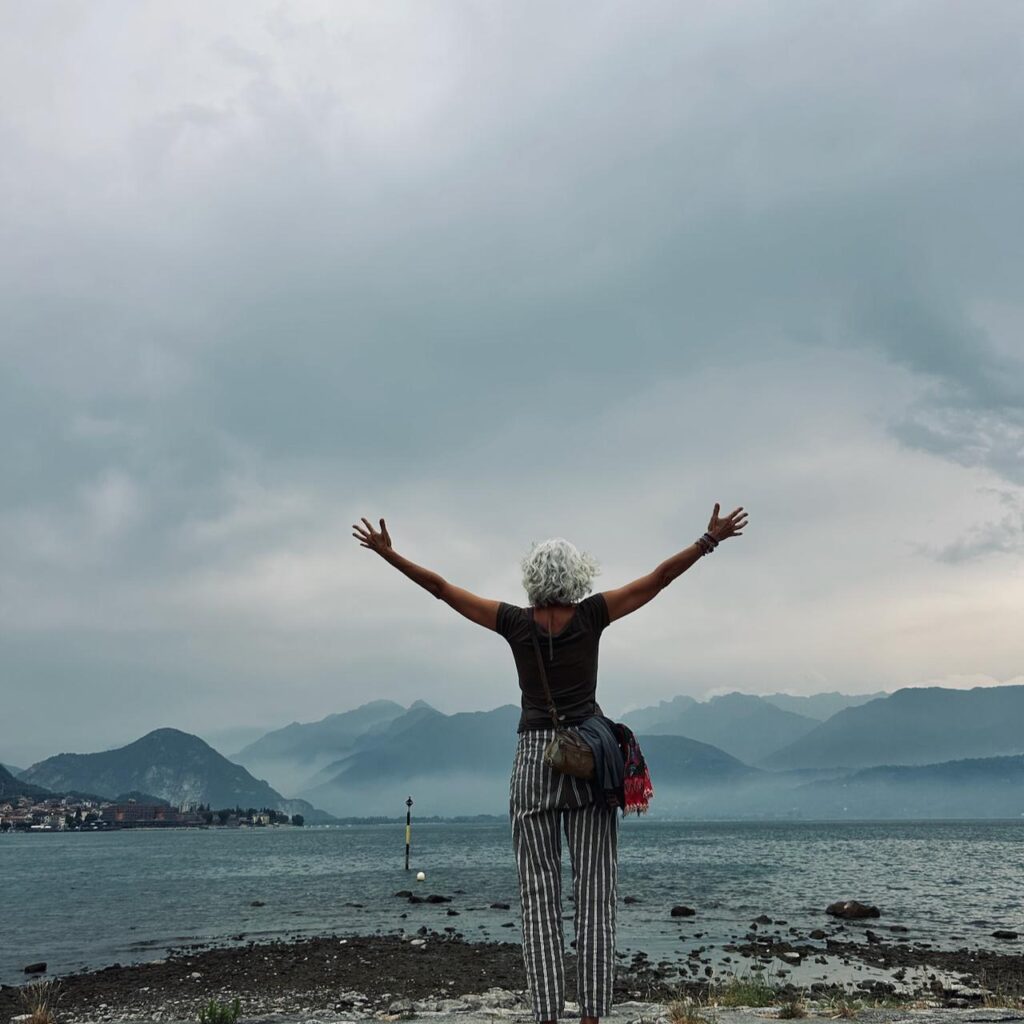 Eine Person mit grauen Haaren steht mit erhobenen Armen an einem felsigen Ufer und blickt unter einem bewölkten Himmel auf einen See und die fernen Berge.