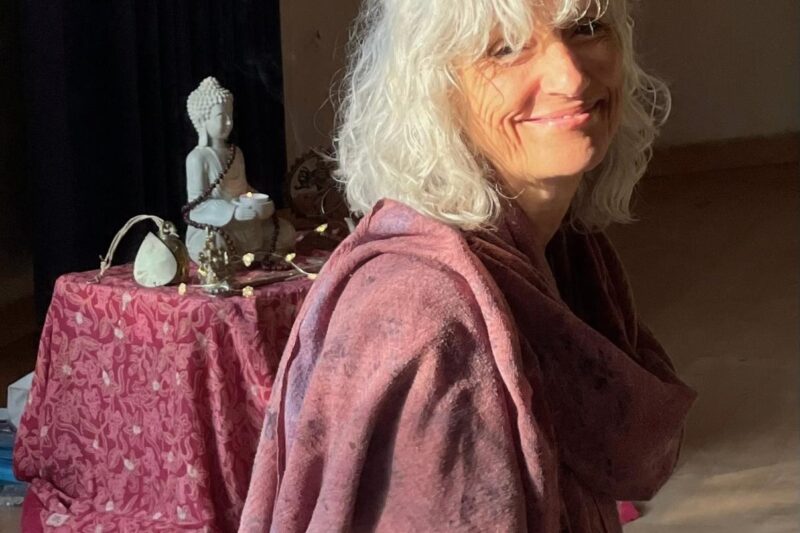 Eine Frau mit weißem Haar lächelt im Sonnenlicht. Sie ist in einen violetten Schal gehüllt. Hinter ihr steht ein Tisch, auf dem eine Buddha-Statue, eine Trommel und weitere Dekorationsgegenstände auf einem gemusterten Tuch stehen.