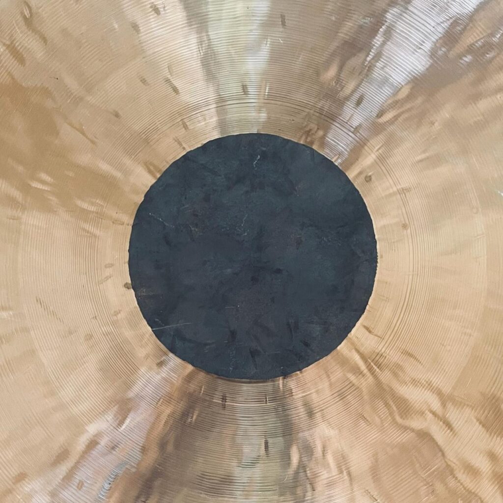 Nahaufnahme eines Gongs mit einer glänzenden, goldenen Oberfläche und einem großen, dunklen, kreisförmigen Zentrum. Lichtreflexe erzeugen einen welligen, strukturierten Effekt auf dem Metall.